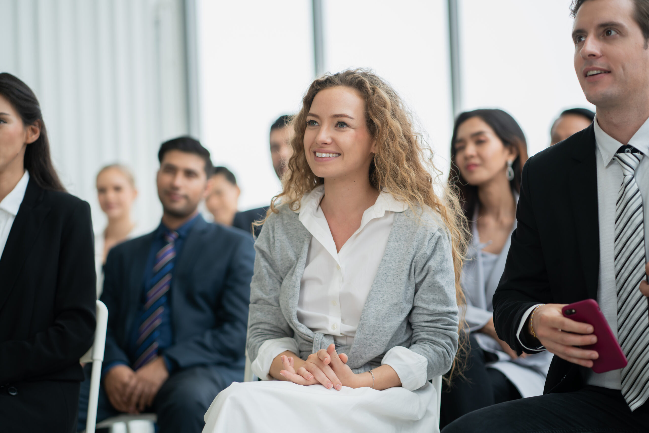 Eine junge Frau mit lockigem Haar sitzt lächelnd im Publikum einer Veranstaltung oder eines Seminars. Um sie herum sitzen weitere Teilnehmer in Business-Kleidung.
