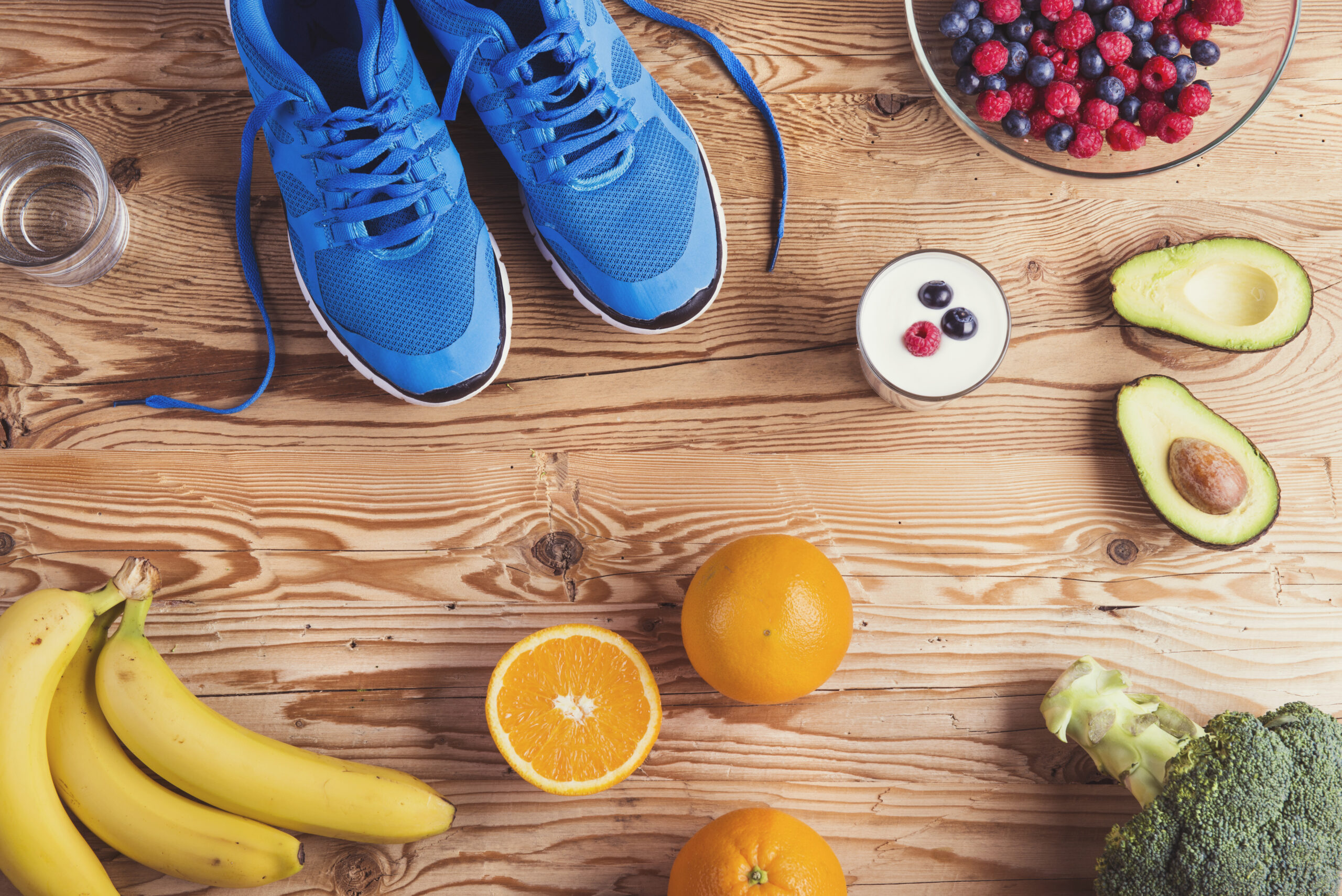 Draufsicht auf einen Holztisch mit Sport- und Ernährungselementen: Ein Paar blaue Laufschuhe, ein Glas Wasser, eine Schale mit Beeren, Joghurt, eine aufgeschnittene Avocado, Orangen, Bananen und Brokkoli.
